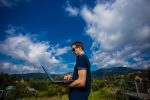 A young man working on a laptop outdoors high in the mountains with a beautiful sky in the background